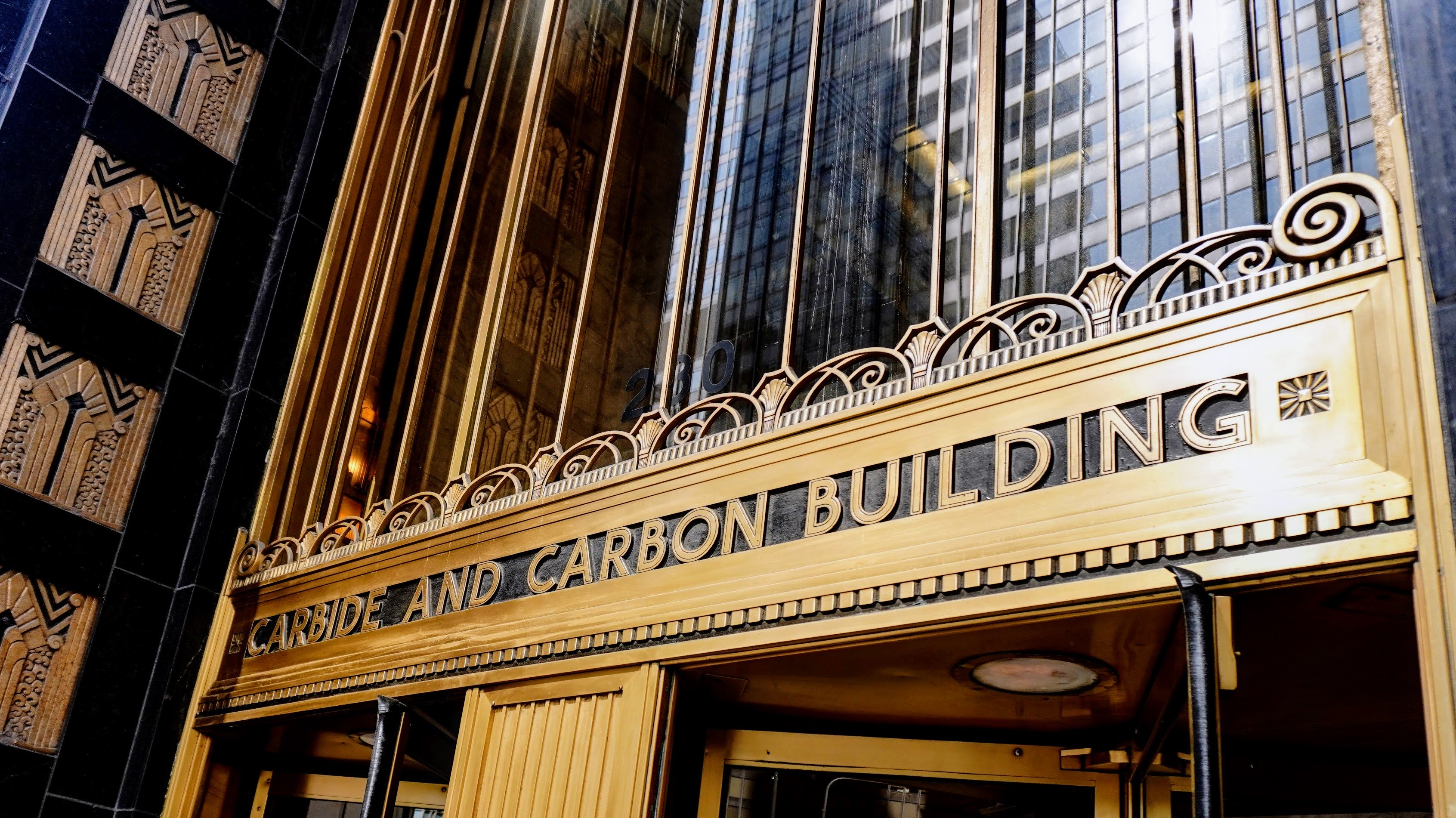 A photo of the Michigan Avenue entrance of the Carbide and Carbon Building. The building's name is written in an art deco font. There are ornamental brass decorations of art deco designs on the left side of the frame.
