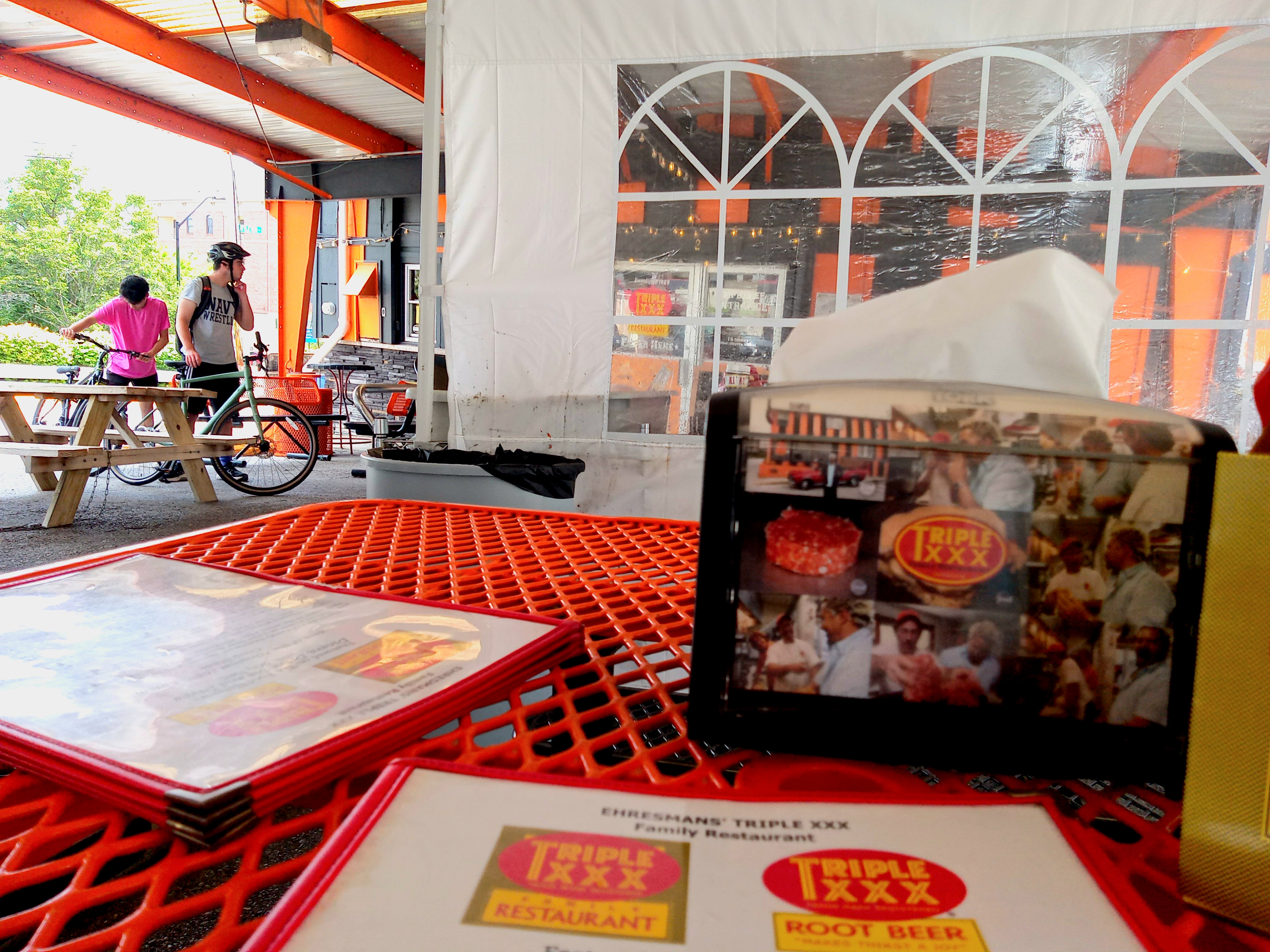 Bikers arrive at Triple XXX Family Restaurant. A view from an outdoor table with a tent to protect from the weather can be seen. The table is orange. There are menus on the table. A view of the front doors and the black and orange walls as well as the ceiling beams can be seen.