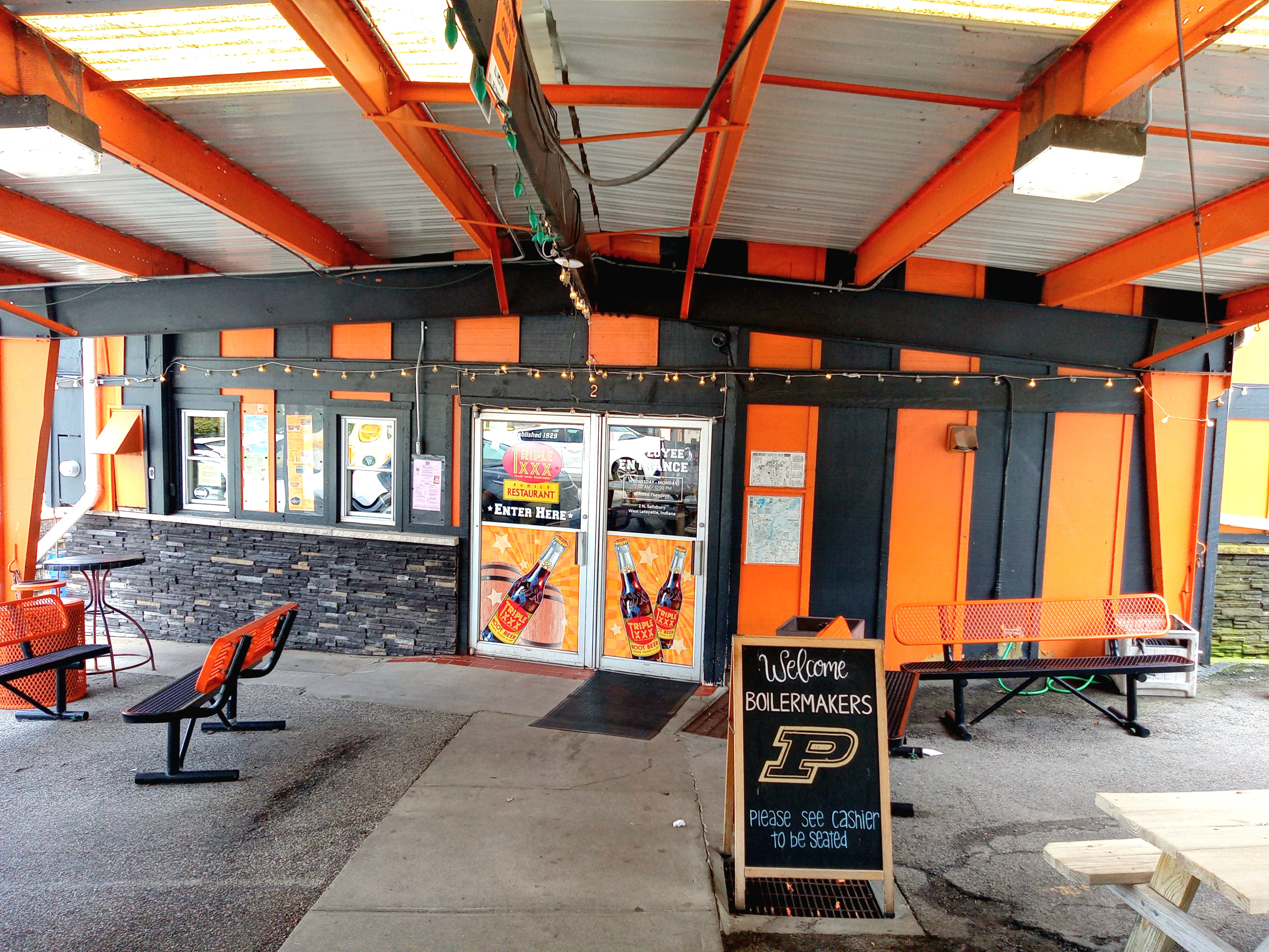 The entrance to Triple XXX Family Restaurant in West Lafayette, Indiana. There are pictures of root beer bottles, the restaurant's logo, and menus tacked to the wall. The colors of the exterior walls are black and orange.